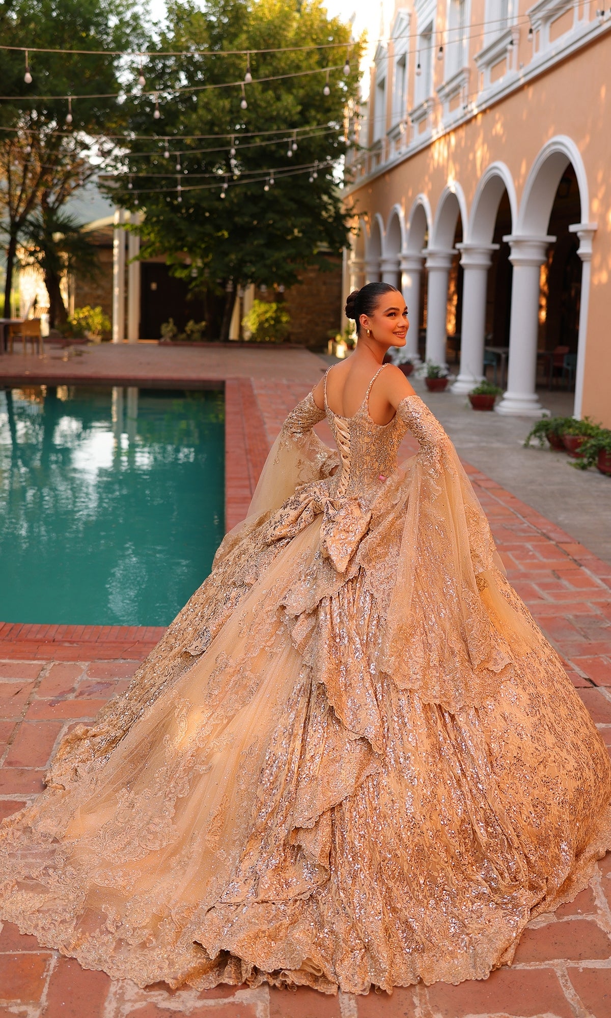 Wearing the Quinceanera Dress 54303 By Amarra, a woman stands beside a pool on a brick patio, framed by trees and an arched building in the background.